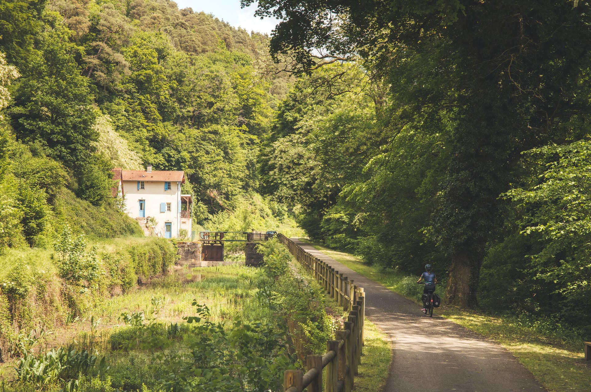 Vue large de la vallée des Éclusiers, véloroute bordée de verdure passant devant une maison éclusière